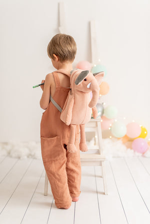 Child wearing a brown romper with a pink plush backpack on a white floor.