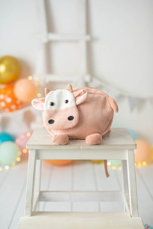 Pink cow backpack on a small wooden stool with colorful balloons in the background